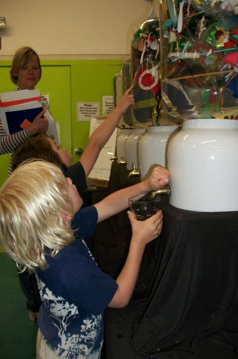 A young boy eagerly fills his glass cup from the fourth water cooler, which jam packed with colorful jetsam representing the amount of plastic pollution in our oceans in 2030, as a concerned parent looks on.