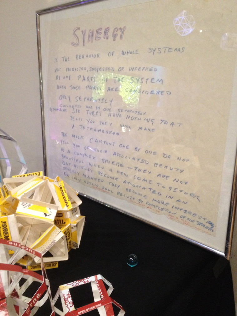 A blue marble sits on the black tablecloth along with a collection of spheres, each constructed from a series of units made from sliced milk cartons. Leaning against the wall is Buckminster Fuller's definition of 'Synergy,' handwritten in blue and red crayon on white paper in a silver metal frame.